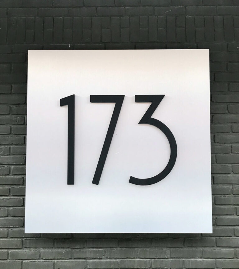 173 Raised Building Address A white sign with the black number 173 mounted on a dark gray brick wall.