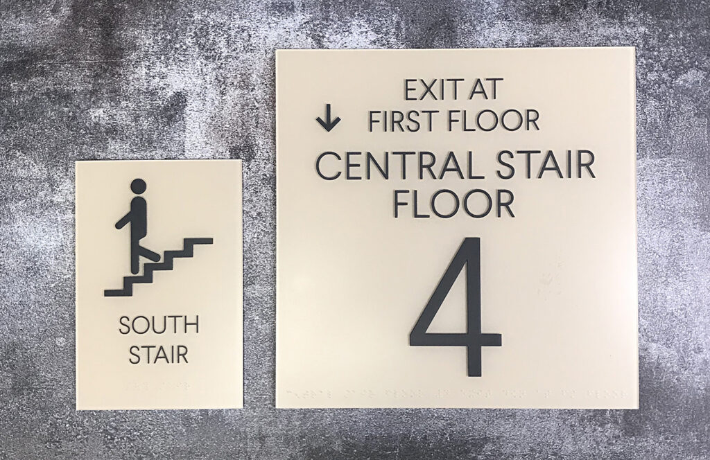 ADA Signage Two signs: one with a stairperson icon and "SOUTH STAIR," another with "EXIT AT FIRST FLOOR, CENTRAL STAIR FLOOR 4.