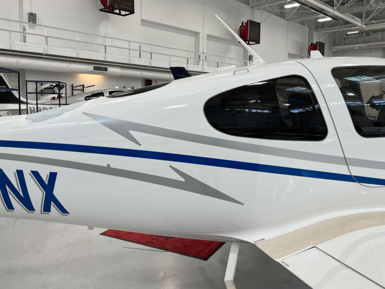 Aircraft Graphics White airplane with blue and gray stripes parked inside a well-lit hangar; partial view of registration letters shown.