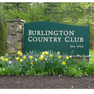 Burlington Country Club Green sign reading "Burlington Country Club Est. 1924" with yellow flowers and greenery in front.