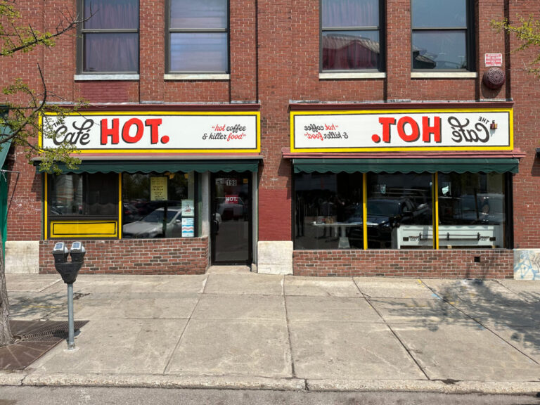 Cafe Hot Custom Hand Painted Sign Brick building with two large windows and a sign reading "Cafe HOT." above the entrance on a city sidewalk.