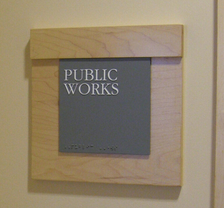 ADA Room Sign A wall-mounted sign with "Public Works" in raised white letters and Braille below on a gray background.