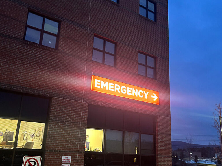 Emergency Sign A brick building with a lit "EMERGENCY" sign and arrow, photographed at dusk.