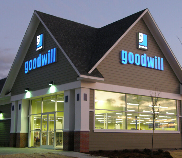 Goodwill Sign Exterior view of a Goodwill store at dusk with illuminated signs and glass entrance doors.