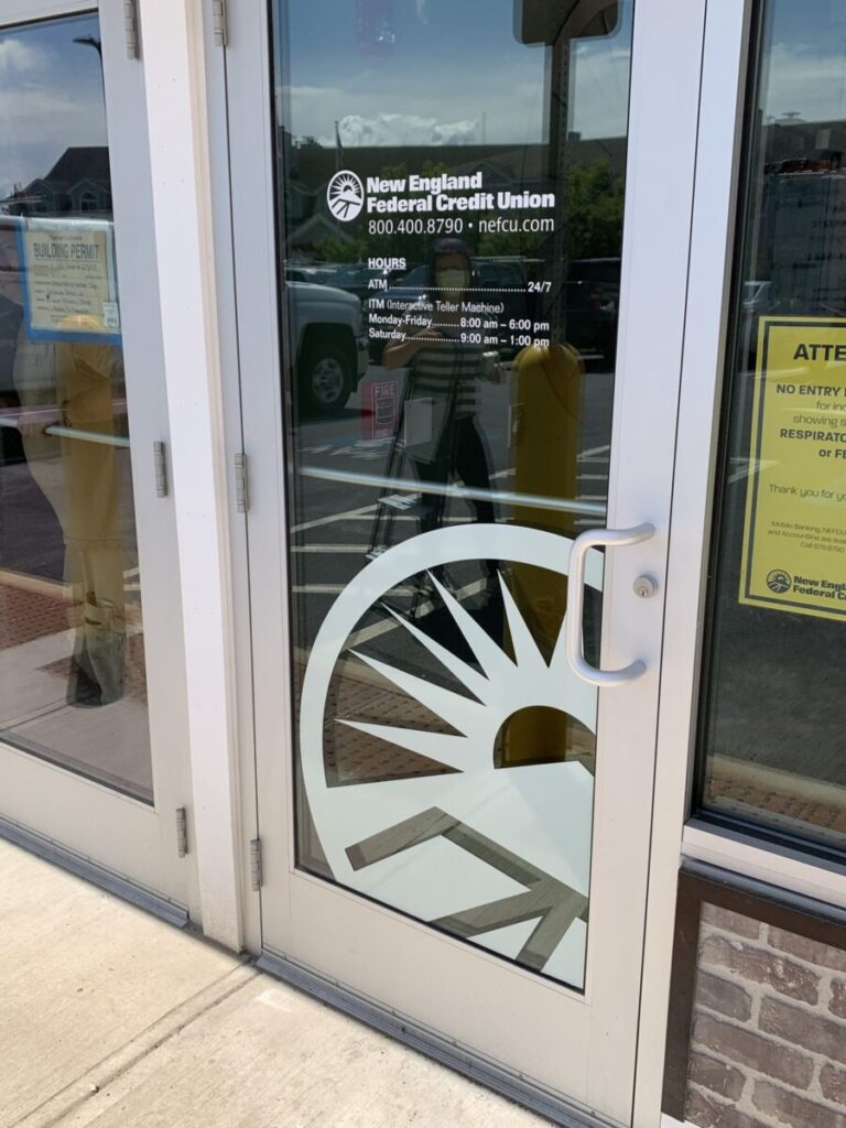 Window Graphics Lettering Glass door of New England Federal Credit Union showing hours, ATM info, and a sunburst logo. Reflections visible on glass.