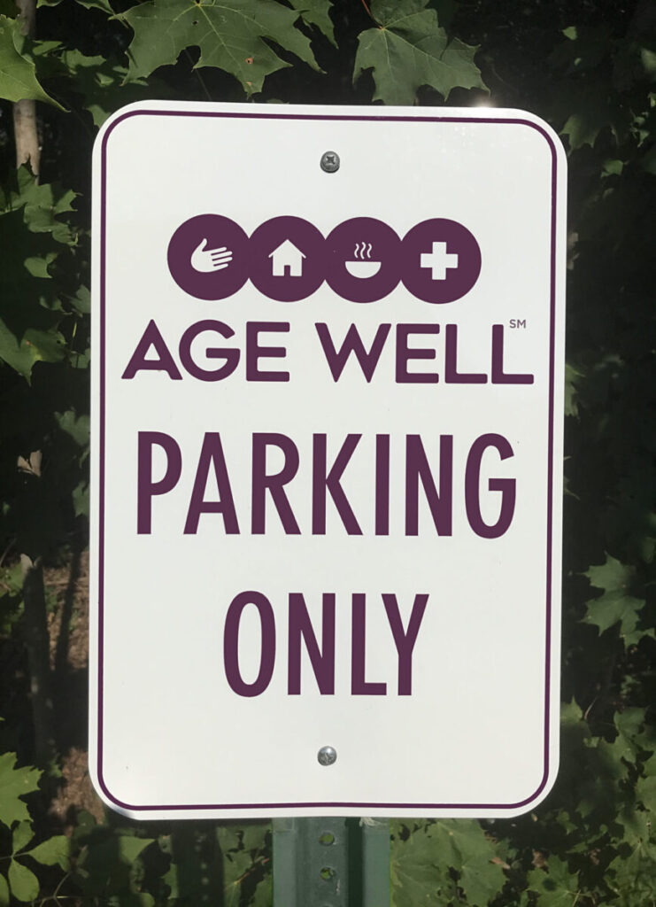 Age Well Parking Signage A sign reading "AGE WELL PARKING ONLY" with icons for care, home, food, and health, posted in front of leafy trees.