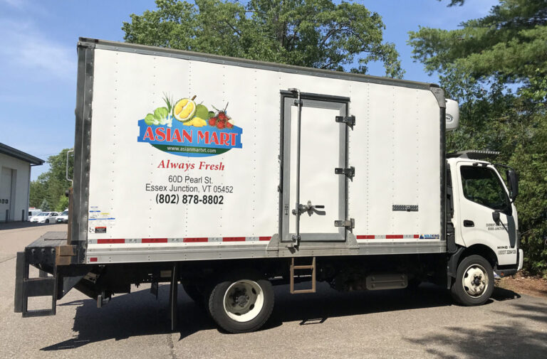 Aisan Mart Truck White delivery truck with "Asian Mart" branding parked on a paved road beside trees on a sunny day.