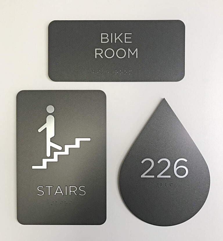 ADA Compliant Signs Three gray signs: "BIKE ROOM," "STAIRS" with a person icon, and a teardrop-shaped "226," each with braille text.