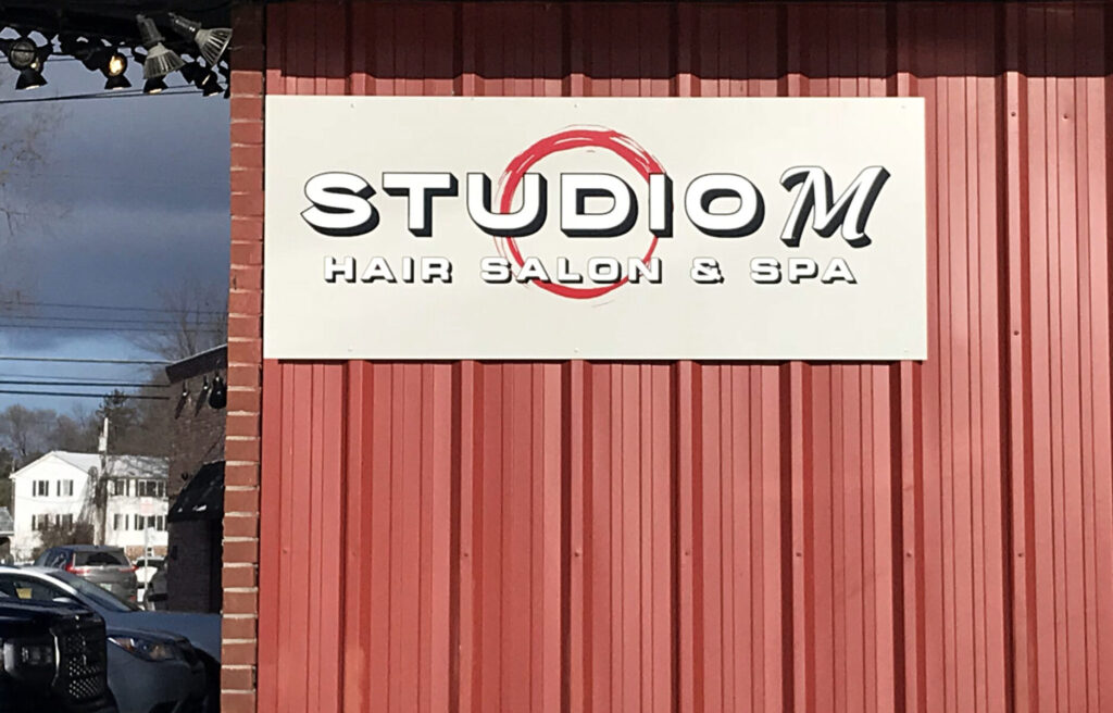 Aluminum Composite Sign Sign reading "STUDIO M HAIR SALON & SPA" mounted on a red corrugated metal wall next to a brick column.