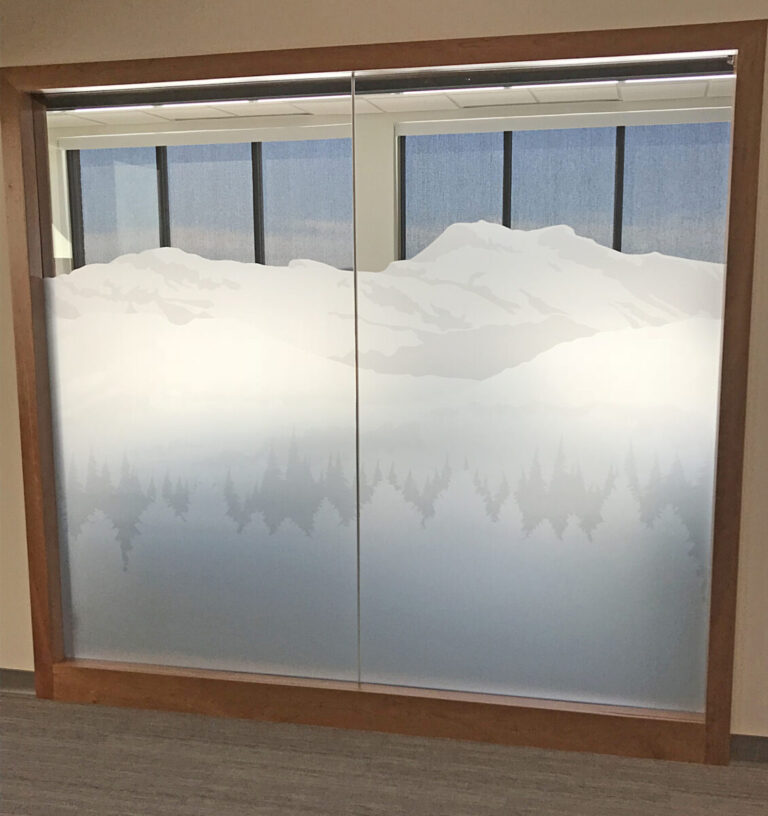 Digital Printed Etched Vinyl Frosted glass window with a mountain and pine tree design, framed by wood, in an indoor office setting.