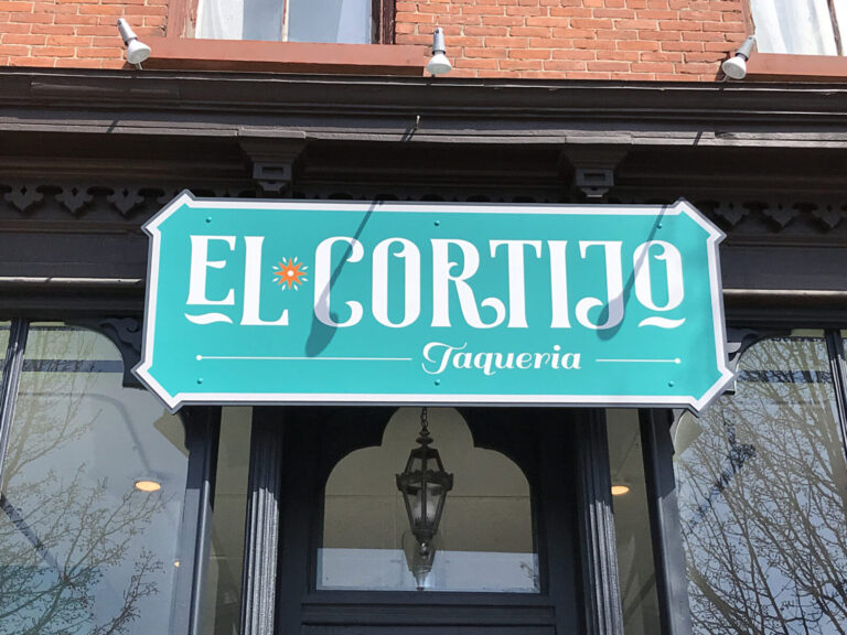 Aluminum Green and white sign reading "El Cortijo Taqueria" mounted above the entrance of a brick building.