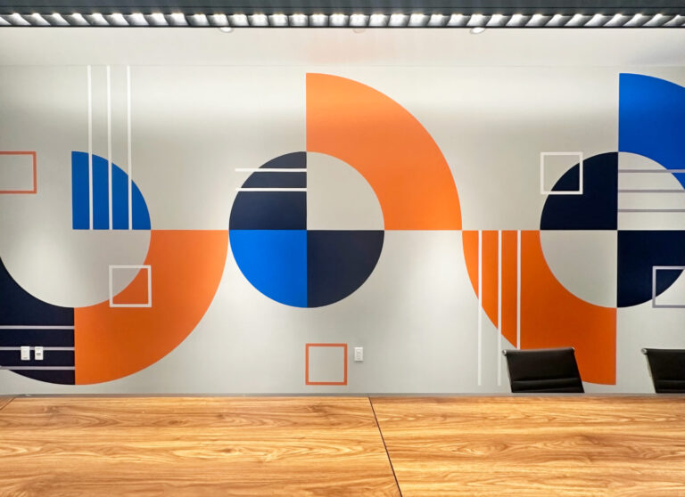 Onlogic Digital Printed Wall Graphic Conference room wall with geometric mural in orange, blue, black, and white patterns, and two wooden tables in front.