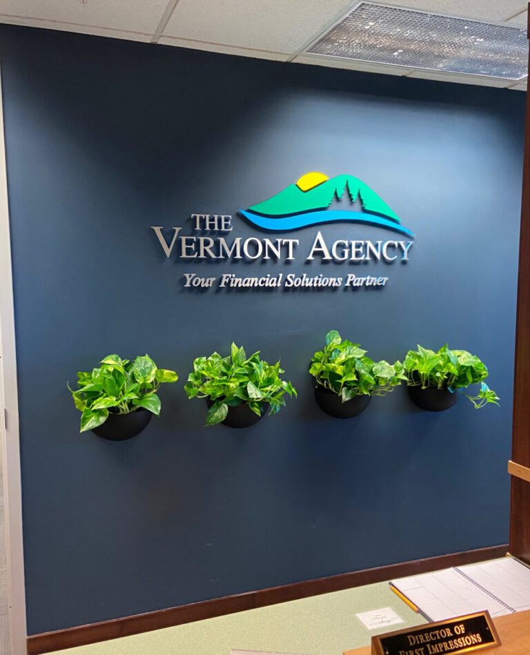 Vermont Agency Raised Wall Graphic Three potted plants on a blue wall beneath "The Vermont Agency" sign with the slogan "Your Financial Solutions Partner.