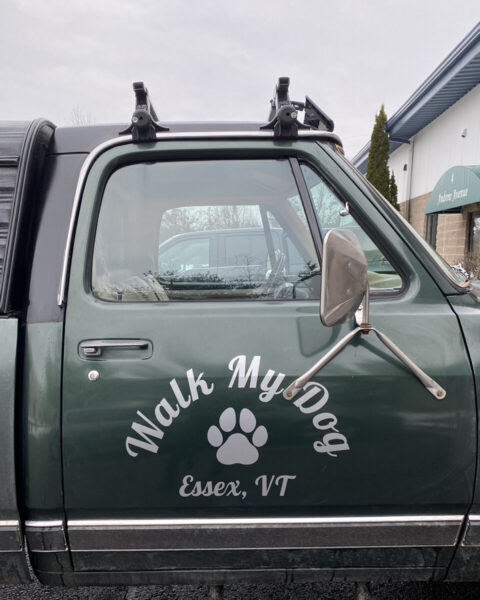 Walk My Dog Green truck door with "Walk My Dog Essex, VT" and a paw print decal on the side, parked near a building.