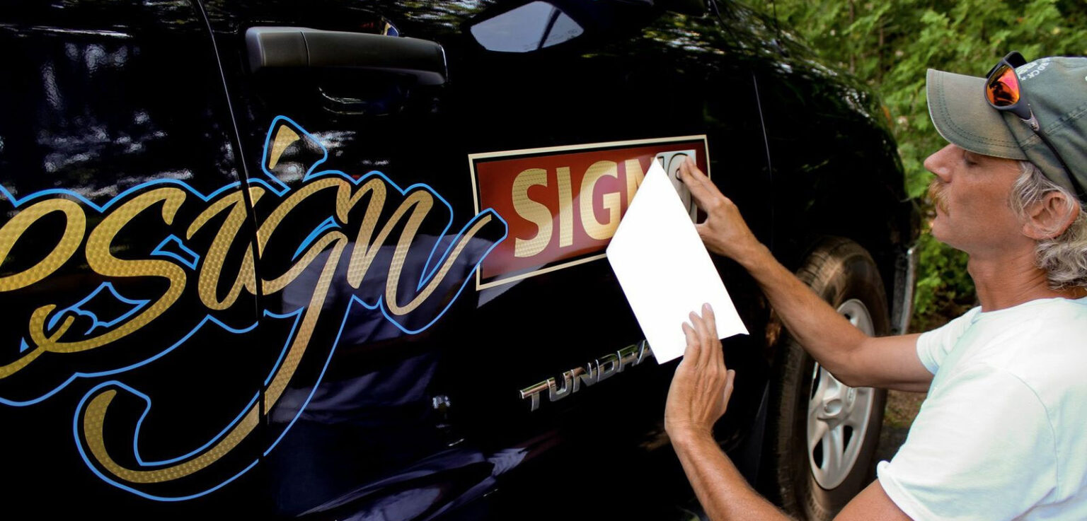 Auto Application A man applies a vinyl sign decal to the side of a black Tundra vehicle while holding a sheet of paper.