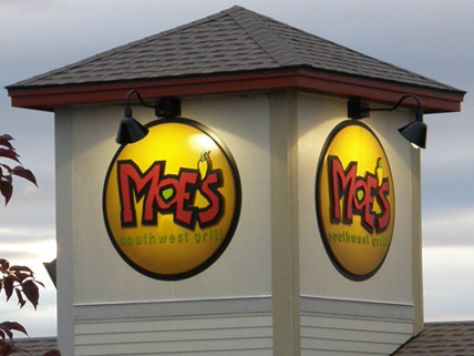 Moe's A building corner displays two illuminated Moe's Southwest Grill signs under exterior lights.