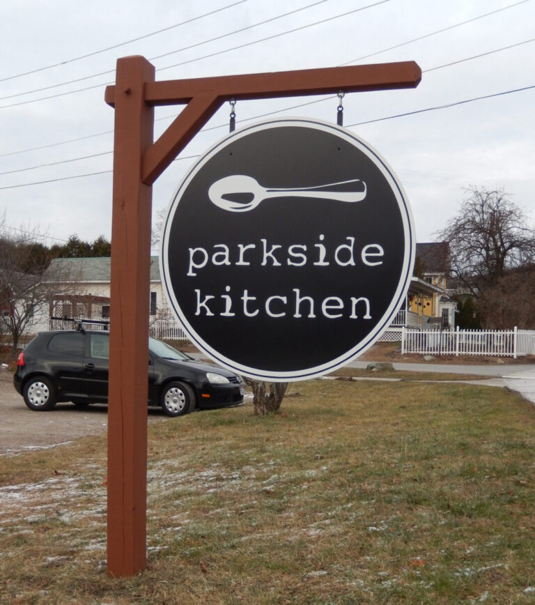 Parkside Kitchen Vinyl Graphic A round sign reading "parkside kitchen" with a spoon graphic, hanging on a wooden post near a grassy area and street.